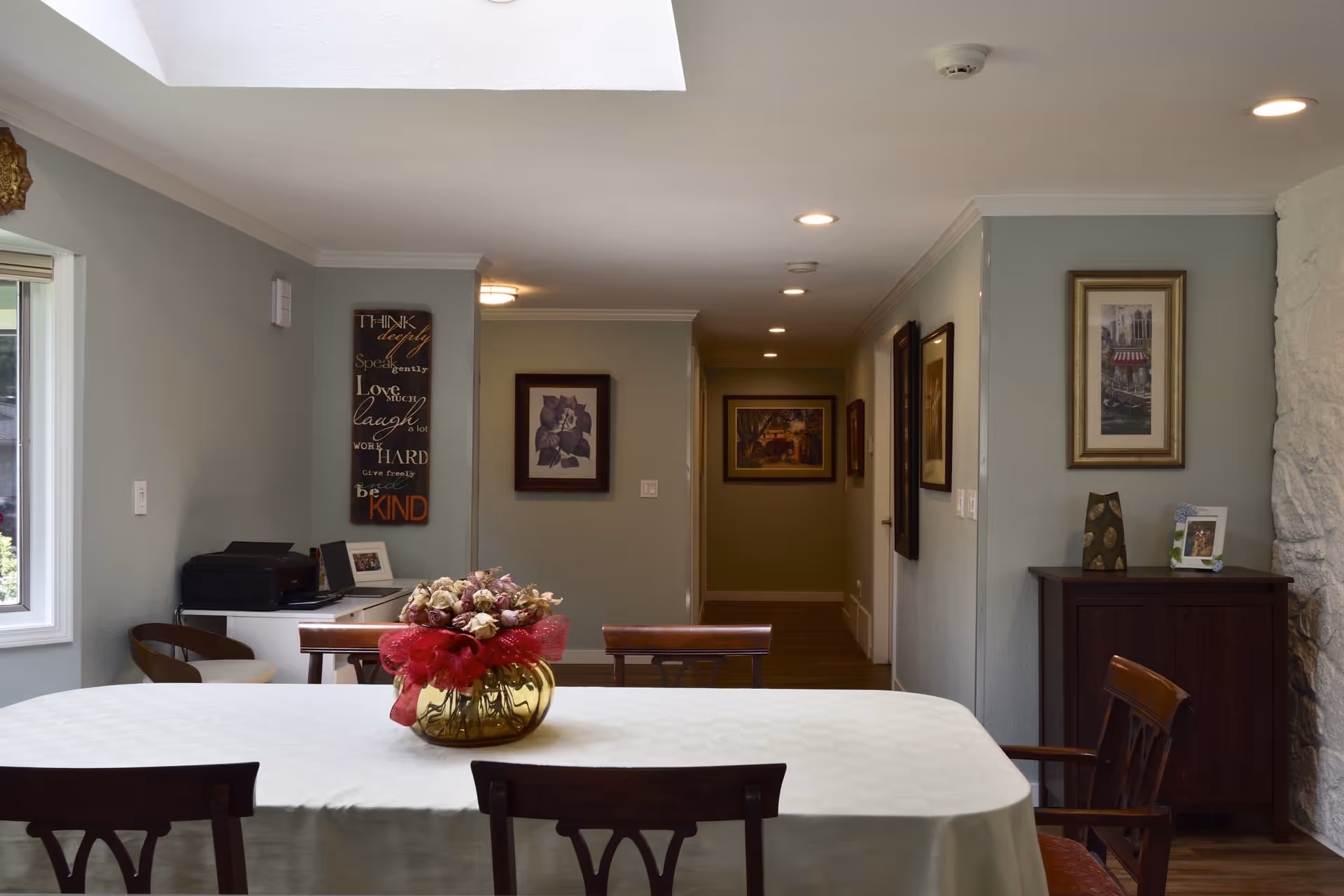 A dining room with a rectangular table covered with a white tablecloth and a centerpiece of dried flowers in a yellow vase with a red ribbon. The room has light green walls adorned with framed artwork and a decorative sign. There is a window on the left side letting in natural light, and a wooden cabinet on the right side with a small framed photo and decorative items. The ceiling has recessed lighting and a skylight.