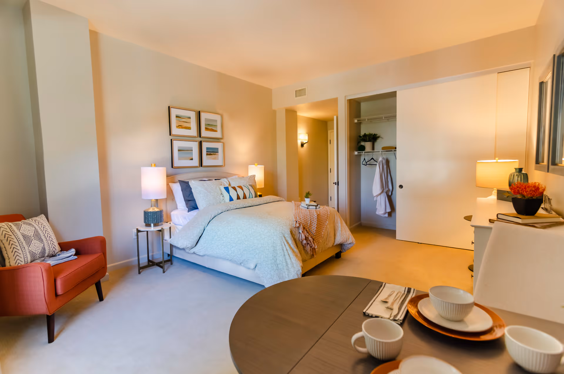 A cozy bedroom in a senior living facility featuring a neatly made bed with pillows and a throw blanket, two bedside tables with lamps, a red armchair with a patterned pillow, an open closet with hanging robes and shelves, and a small dining table set with plates, bowls, and cups. The room is warmly lit with soft yellow lighting.
