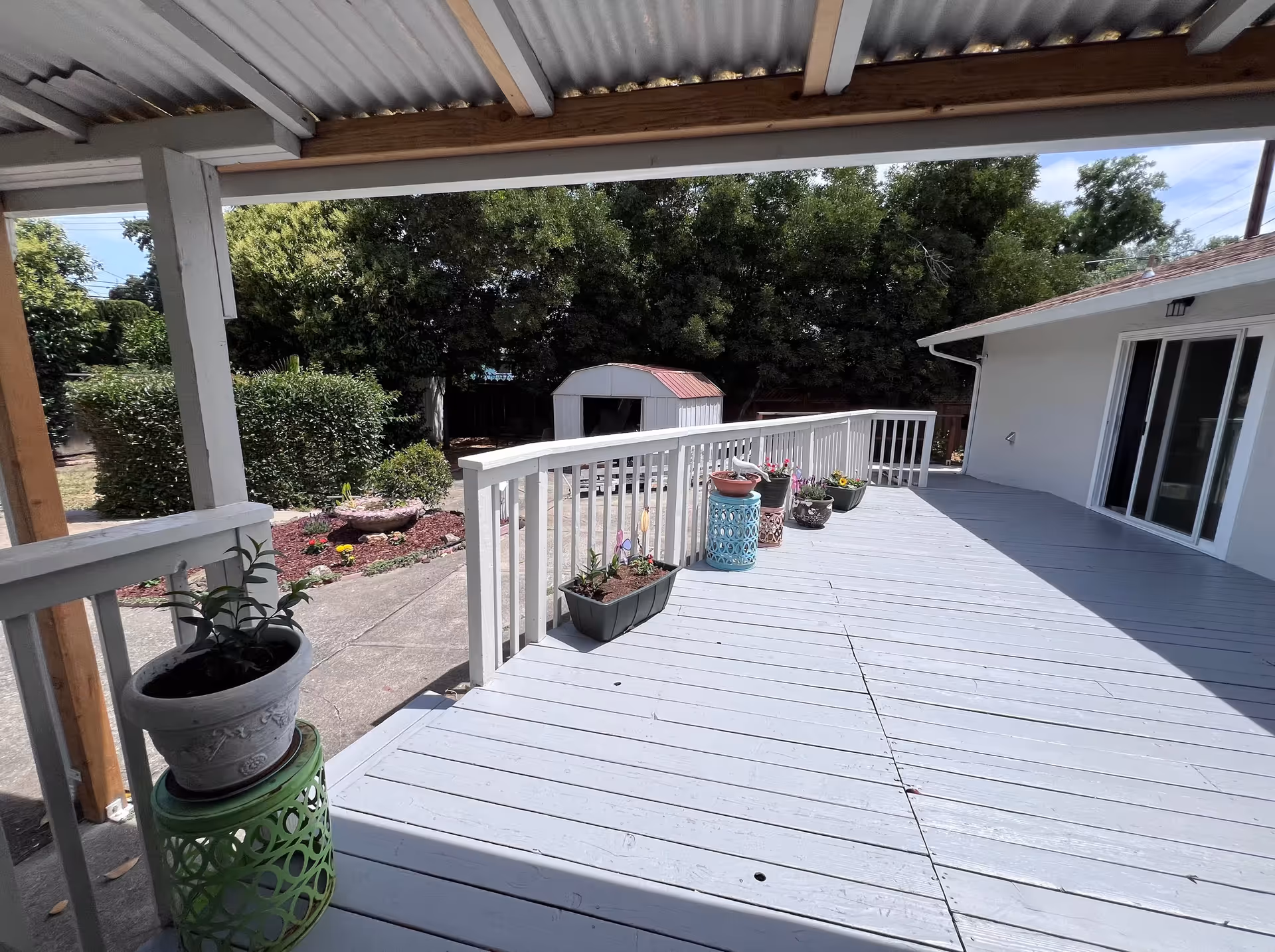 Covered wooden deck with potted plants overlooks a backyard and shed beside a sliding door.
