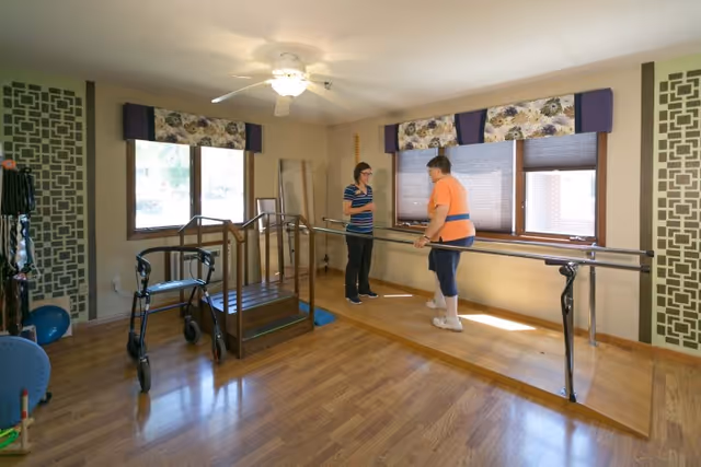 A therapy room with wooden flooring and two windows with floral valances. A woman in an orange shirt is using parallel bars for walking practice, assisted by another woman in a striped shirt. The room contains mobility aids including a walker and a set of stairs with handrails. The walls have decorative panels and there is a ceiling fan with a light.
