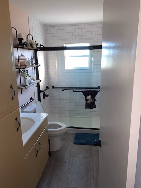 Bathroom with a vanity and sink on the left, toilet, and a tiled walk-in shower with sliding glass doors, grab bars, and a small window.