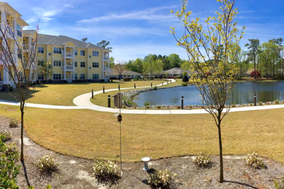A landscaped outdoor area at Porters Neck Village featuring a curved walking path around a small pond, young trees, shrubs, and a three-story yellow residential building in the background under a clear blue sky.