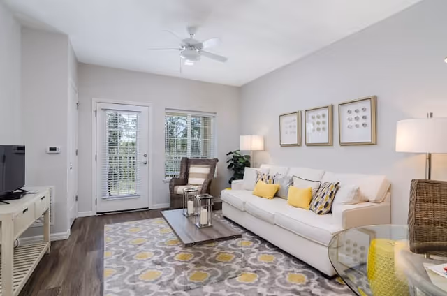 A bright living room featuring a white sofa with yellow and patterned cushions, a wooden coffee table with decorative lanterns, a patterned area rug with yellow and gray tones, a wicker armchair with a cushion, a glass side table with a yellow ottoman underneath, two floor lamps, three framed artworks on the wall, and a door with windows leading outside.