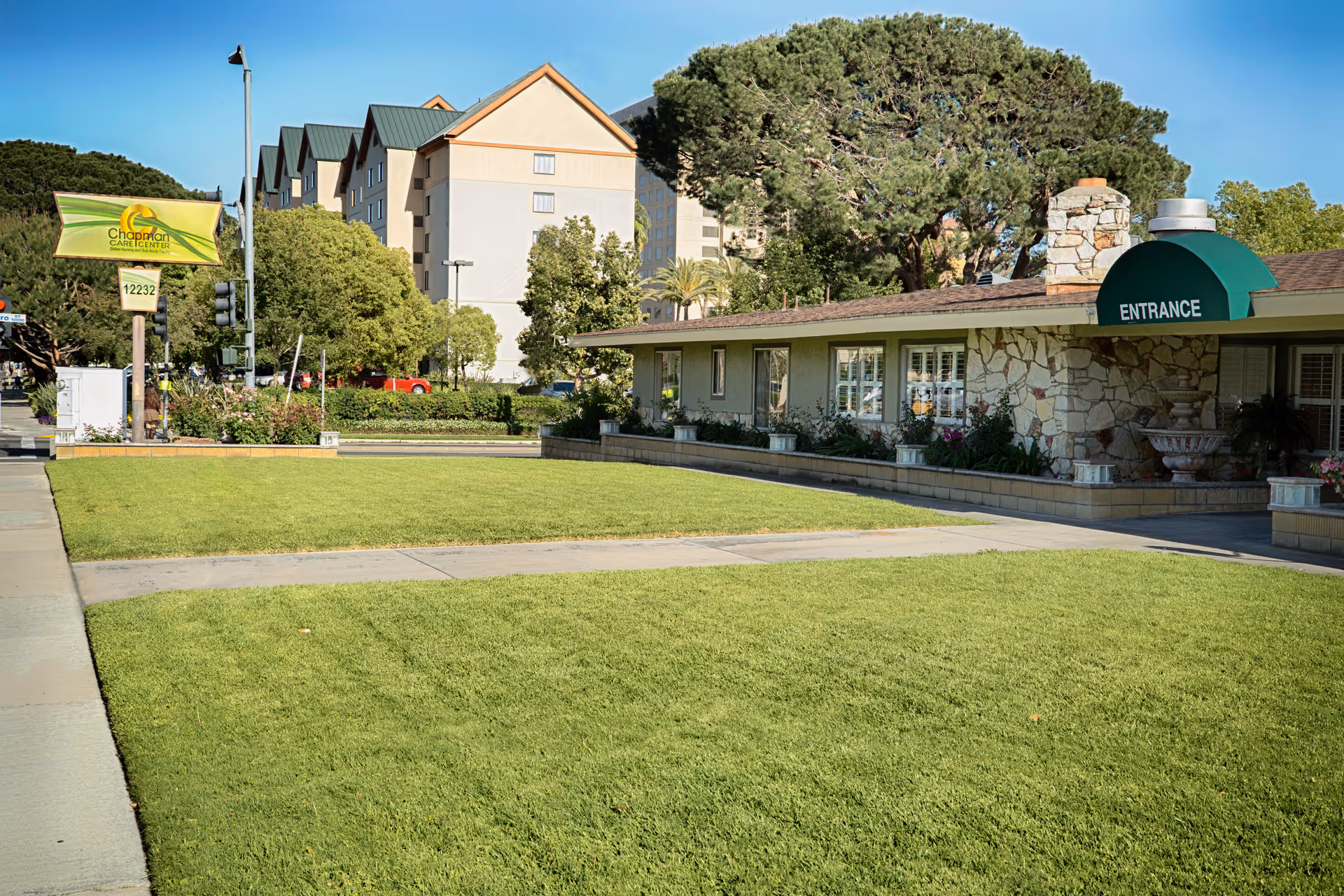 Exterior view of Chapman Care Center showing a well-maintained lawn, a stone building with an entrance marked by a green awning, and a sign with the facility's name and address near the sidewalk.
