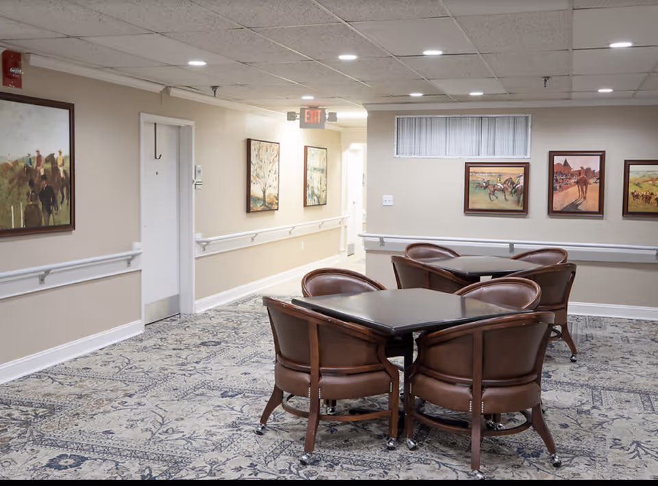 Carpeted communal seating area with two square tables surrounded by leather chairs in a hallway decorated with framed paintings.