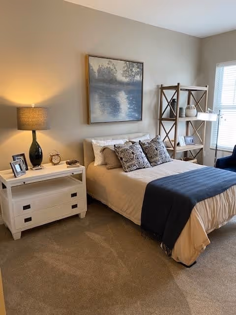 A cozy bedroom with a neatly made bed featuring beige bedding and decorative pillows. A dark blue throw blanket is draped across the foot of the bed. To the left of the bed is a white nightstand with a green lamp, a clock, and framed photos. Above the bed hangs a framed painting of a serene lakeside scene. To the right, there is a wooden shelving unit with decorative items and a window letting in natural light.