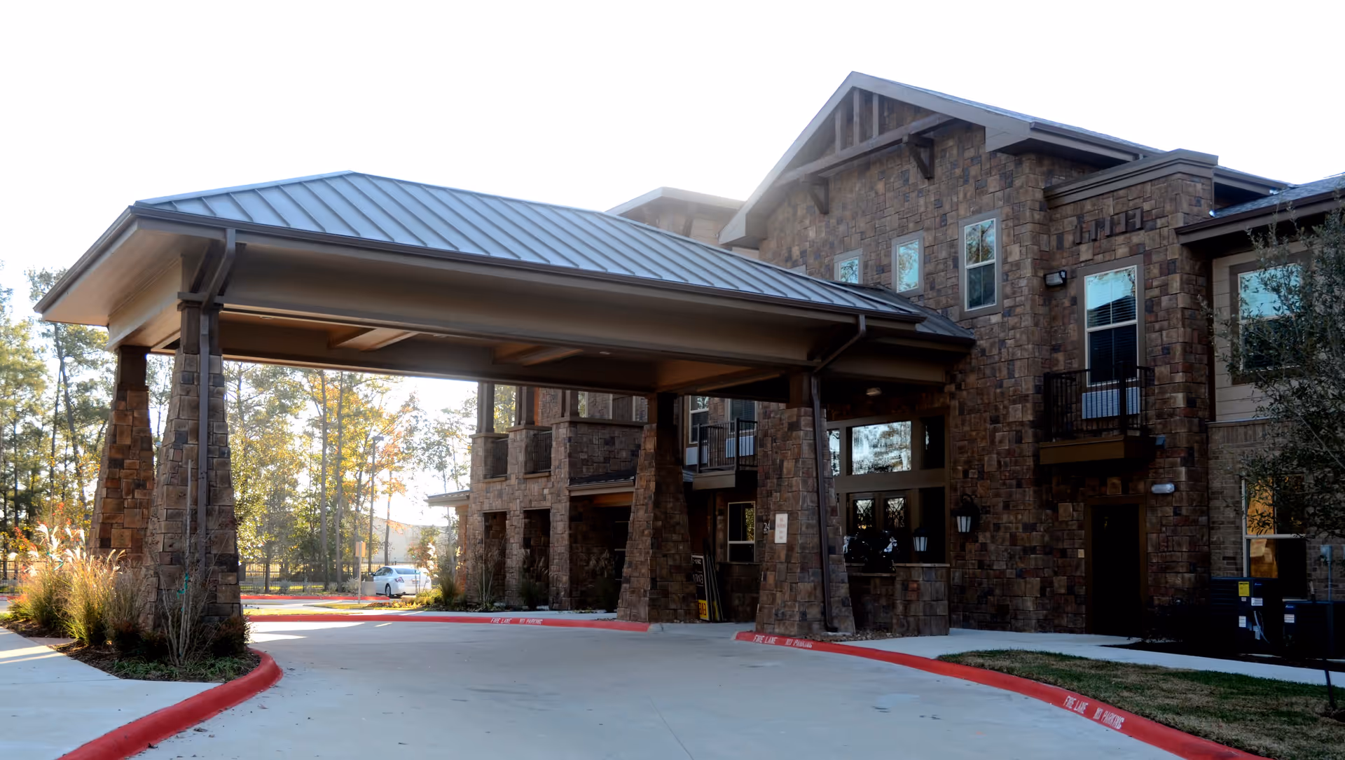 Exterior view of a senior living facility named Landon Ridge Kingwood, showing a covered entrance with stone pillars and a driveway surrounded by trees and landscaping.