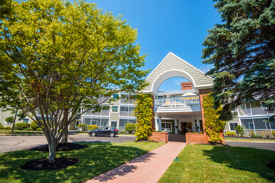 Exterior view of a senior living facility named The Highlands, featuring a brick walkway leading to a covered entrance with a triangular roof and archway. The building has multiple balconies and is surrounded by green trees and well-maintained landscaping under a clear blue sky.