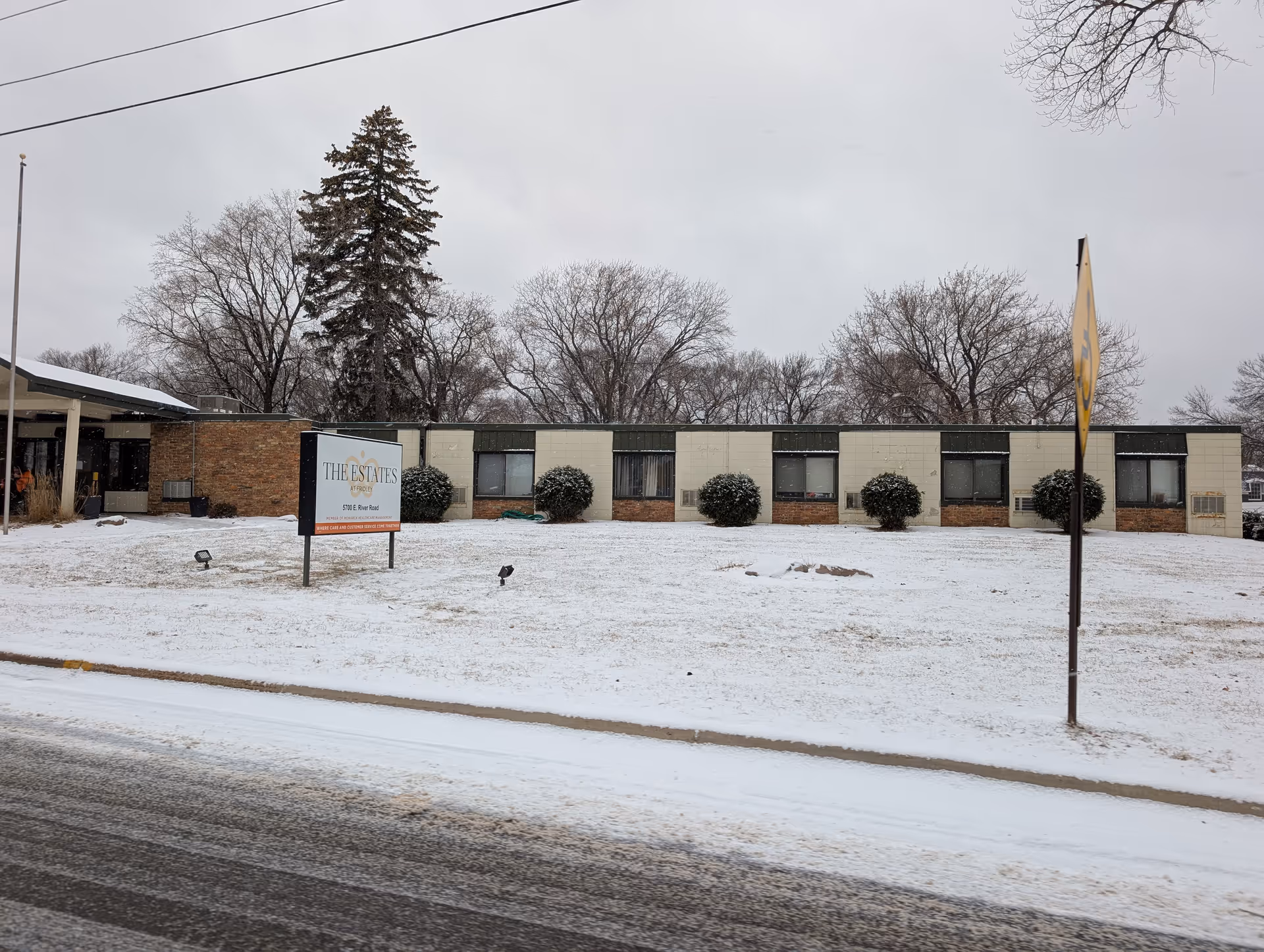 Exterior view of The Estates at Fridley building on a snowy day with a sign in front displaying the facility name and address. The building is single-story with several windows and bushes in front, and leafless trees in the background.