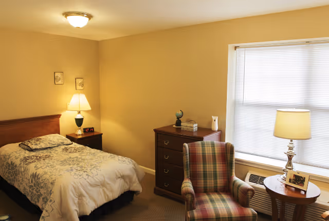 A cozy bedroom with a single bed covered in a floral comforter, a wooden nightstand with a lamp and digital clock, a wooden dresser with decorative items, a plaid upholstered armchair, a round wooden side table with a lamp and framed photo, and a window with closed blinds.