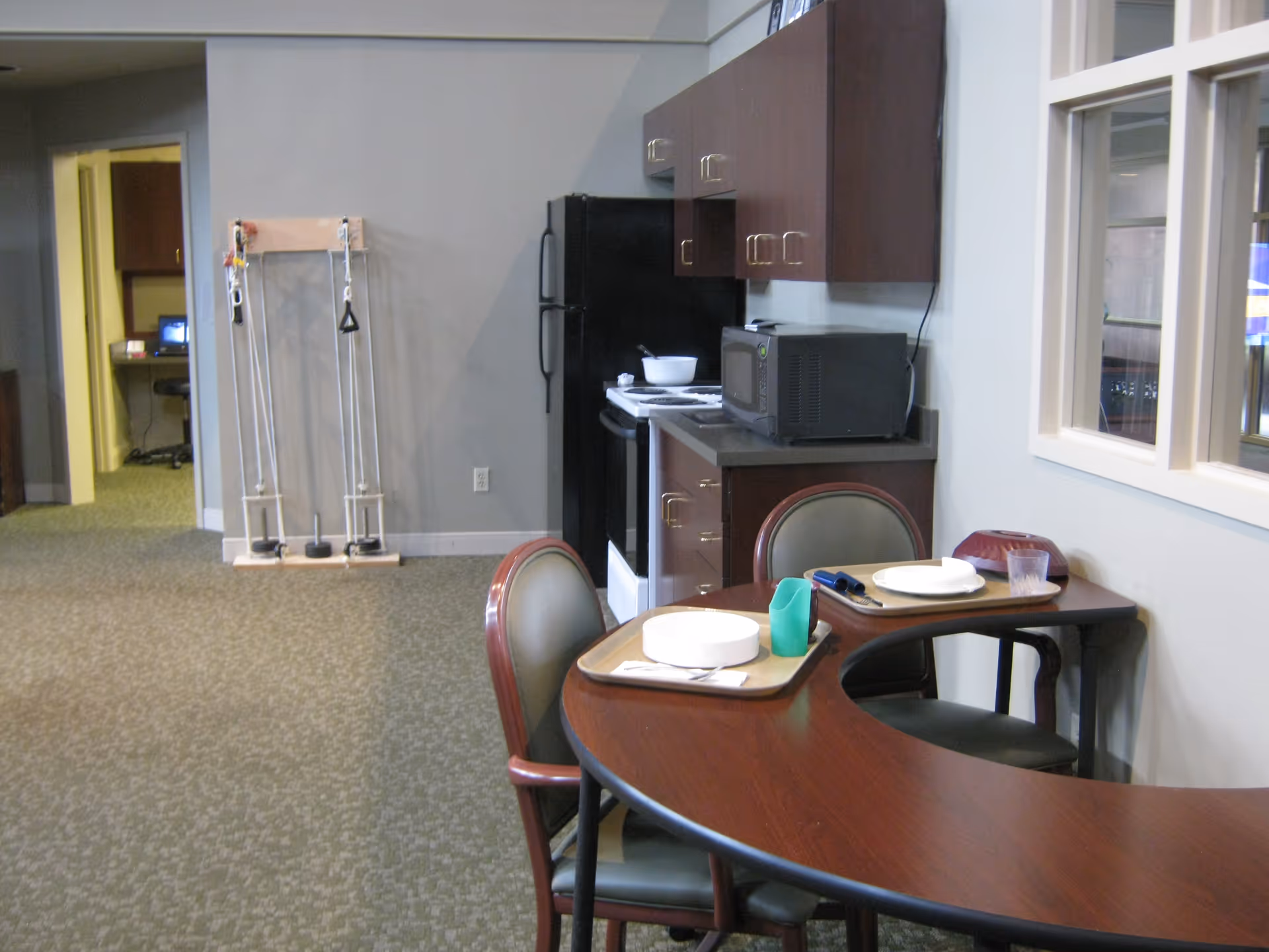 Interior view of a senior living facility kitchen area with a small dining table set for two. The kitchen has dark wood cabinets, a black refrigerator, a microwave, and a stove. There is exercise equipment mounted on the wall in the background and a computer workstation visible through an open doorway.