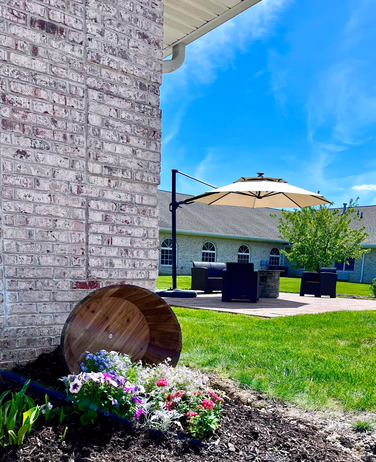 A landscaped courtyard showing an overturned wooden planter with flowers beside a brick wall and a patio seating area with a large umbrella on a grassy lawn.