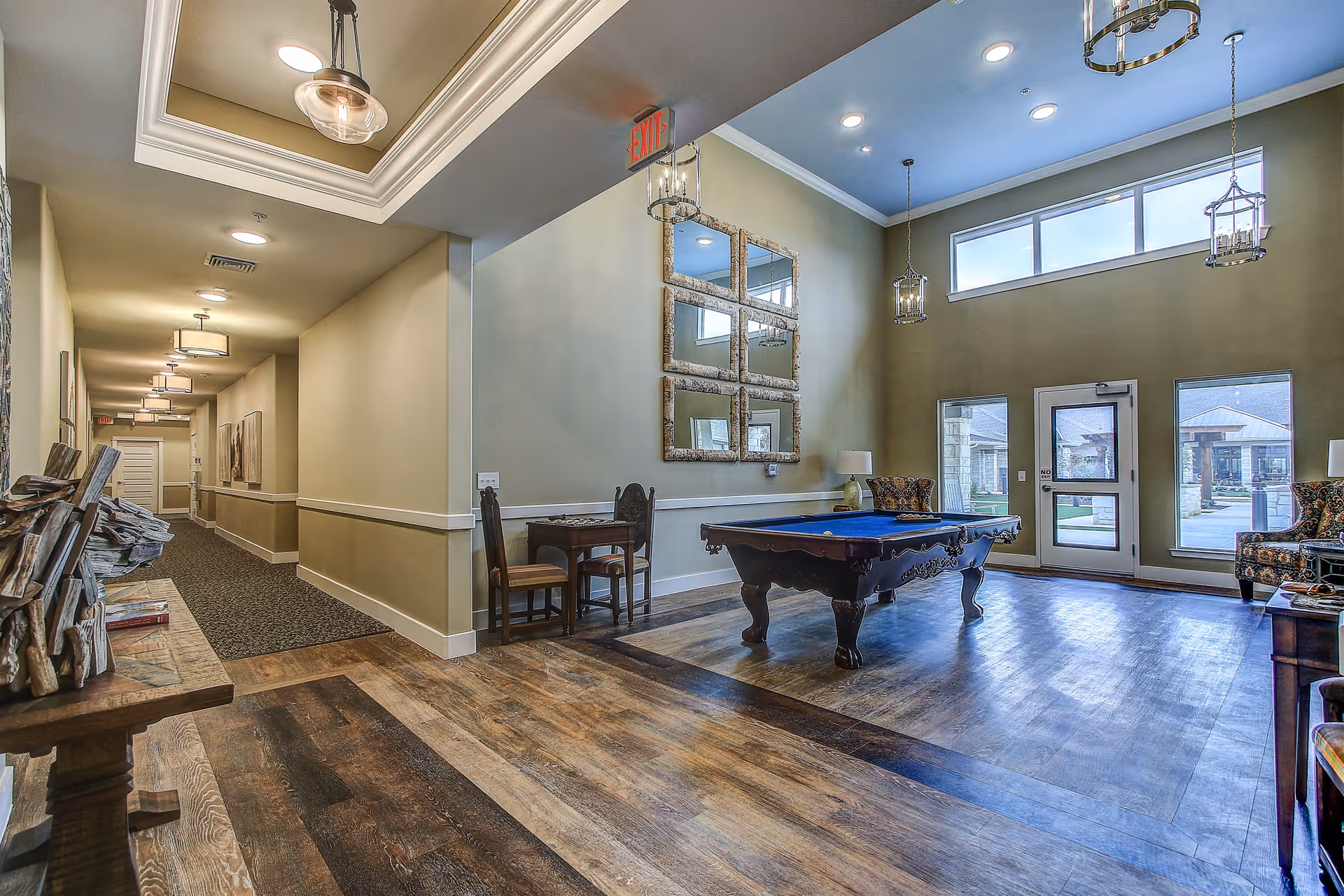 Interior view of a senior living facility common area with a pool table in the center, two armchairs, a small table with two chairs, large windows letting in natural light, and a hallway leading to other rooms. The room has wooden flooring, beige walls, and decorative lighting fixtures.