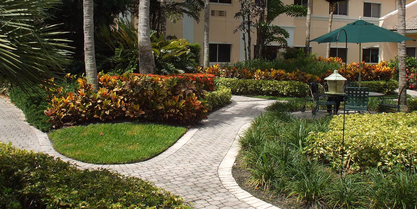 A landscaped outdoor garden area with a winding paved pathway surrounded by green grass, colorful shrubs, and palm trees. There is a seating area with a green umbrella, chairs, and a table near a building with beige walls and windows.