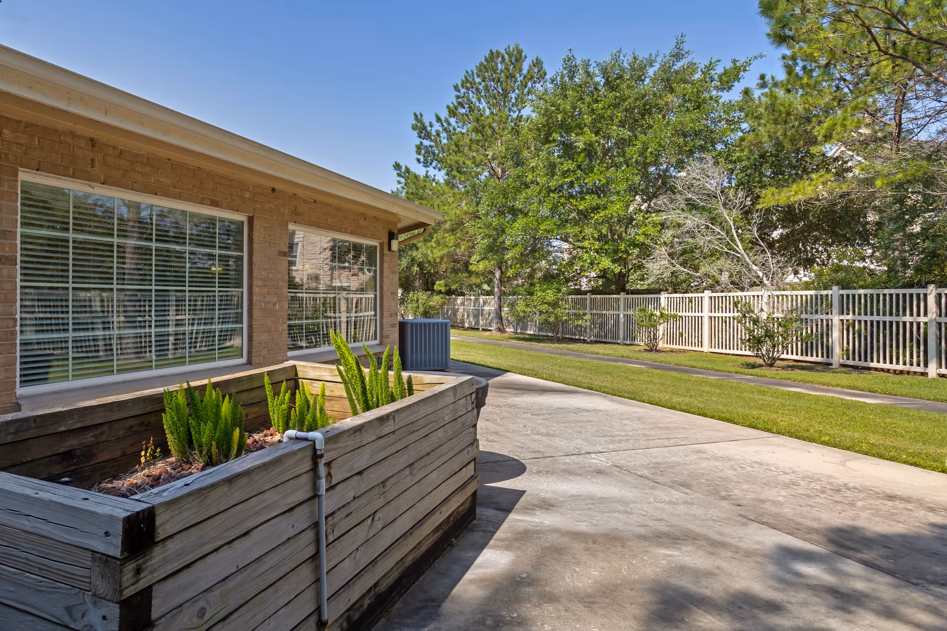 Outdoor area of Cinco Ranch Alzheimer's Special Care Center featuring a wooden raised planter with green plants, a concrete walkway, a brick building with large windows, and a white fence with trees and grass in the background under a clear blue sky.