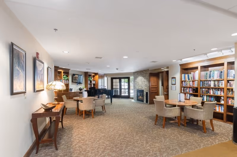 A spacious, well-lit common area in a senior living facility featuring a carpeted floor, several round tables with upholstered chairs, a bookshelf filled with books, a stone fireplace, and a grand piano near large windows and glass doors.