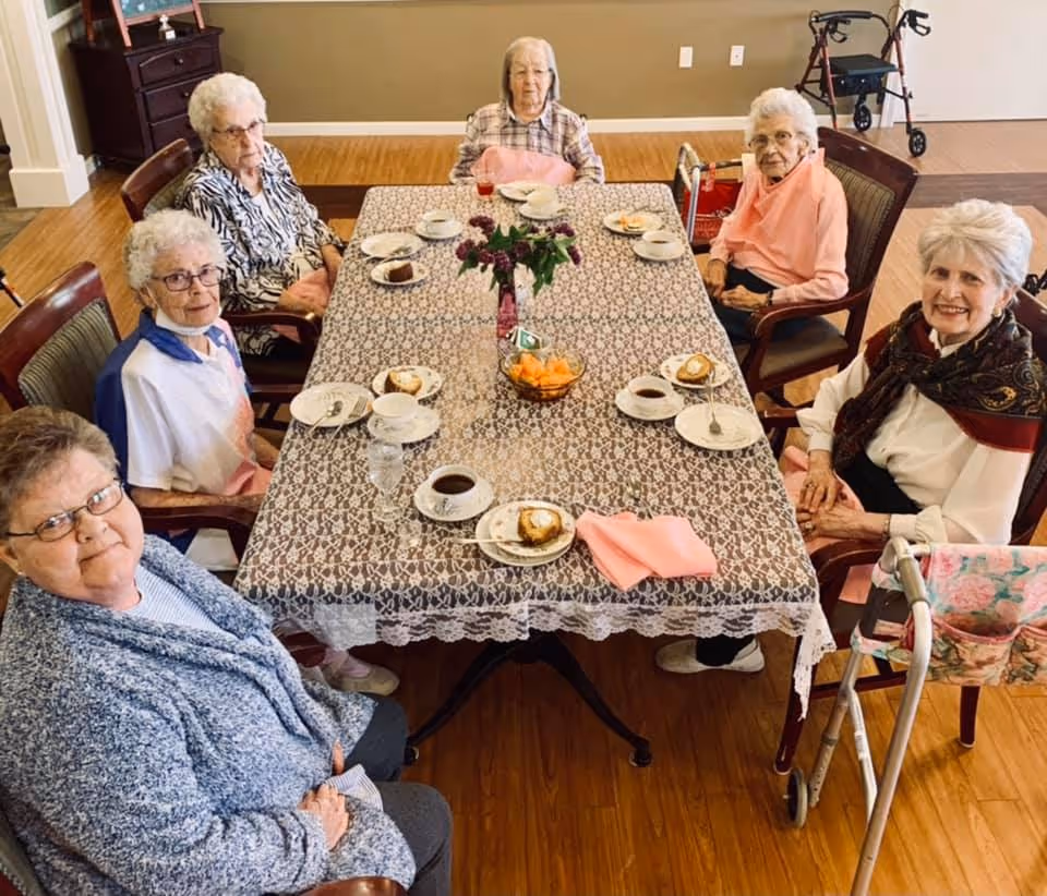 Six elderly women sitting around a rectangular dining table covered with a lace tablecloth. The table is set with plates of dessert, cups of coffee, and a bowl of fruit in the center. The women are seated in a well-lit room with wooden flooring, and a walker is visible near one of the women.