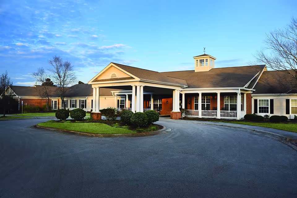 Exterior view of Spring Arbor of Wilmington senior living facility showing a single-story building with a covered entrance supported by white columns, surrounded by neatly trimmed bushes and a circular driveway under a partly cloudy blue sky.