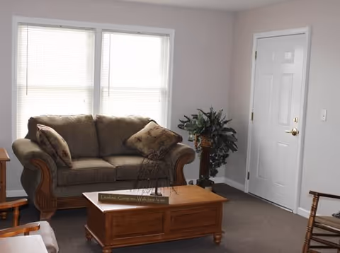 A cozy living room with a brown upholstered sofa adorned with two patterned pillows, a wooden coffee table with a decorative item and a small sign, a potted plant on a stand near a white door, and a window with closed blinds letting in natural light.