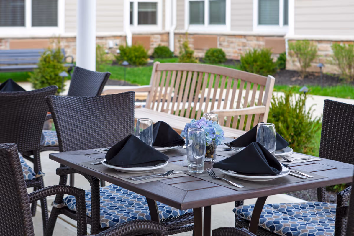 Outdoor patio dining area with a table set for four, featuring black napkins, clear drinking glasses, silverware, and a small vase with purple flowers. The table is surrounded by wicker chairs with patterned cushions, and a wooden bench is visible in the background along with green shrubs and a building exterior.