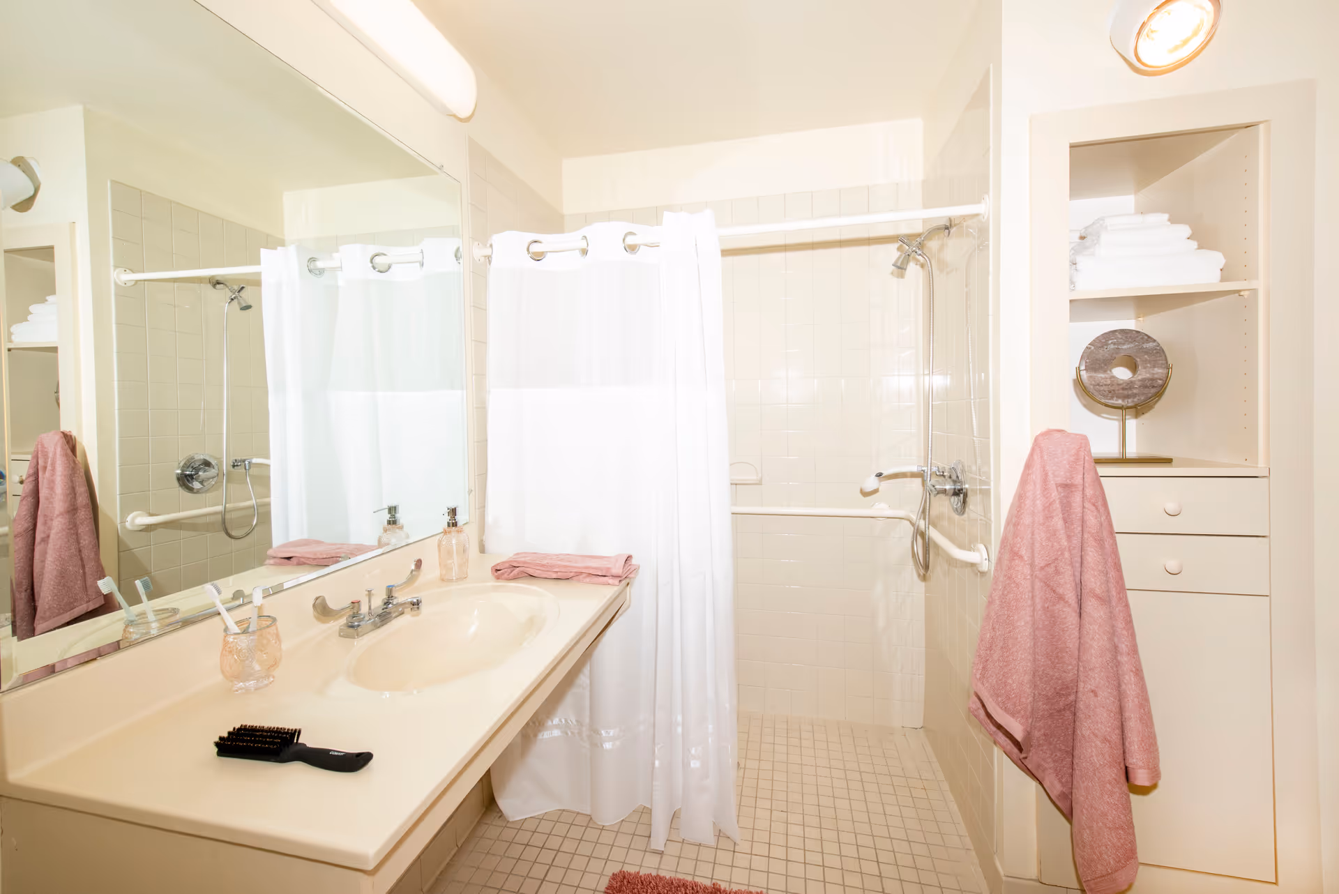 A clean bathroom with a beige countertop and sink, a large mirror above the sink, a white shower curtain enclosing a tiled shower area with a handheld showerhead, pink towels hanging on a rack and folded on the counter, and shelves with neatly stacked white towels and a decorative item.