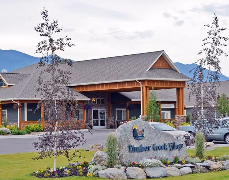Exterior view of Timber Creek Village Community building with a large covered entrance, landscaped garden with flowers and rocks, and a stone sign displaying the community name. Mountains are visible in the background under a cloudy sky.