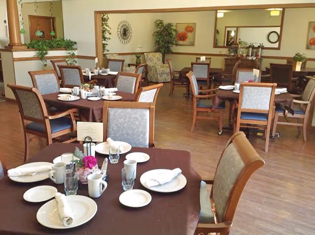 A dining room in a senior living facility with several round tables covered with brown tablecloths. Each table is set with white plates, cups, glasses, and napkins. There are wooden chairs with cushioned seats around the tables. The room has wooden flooring, plants, and framed artwork on the walls, creating a warm and inviting atmosphere.