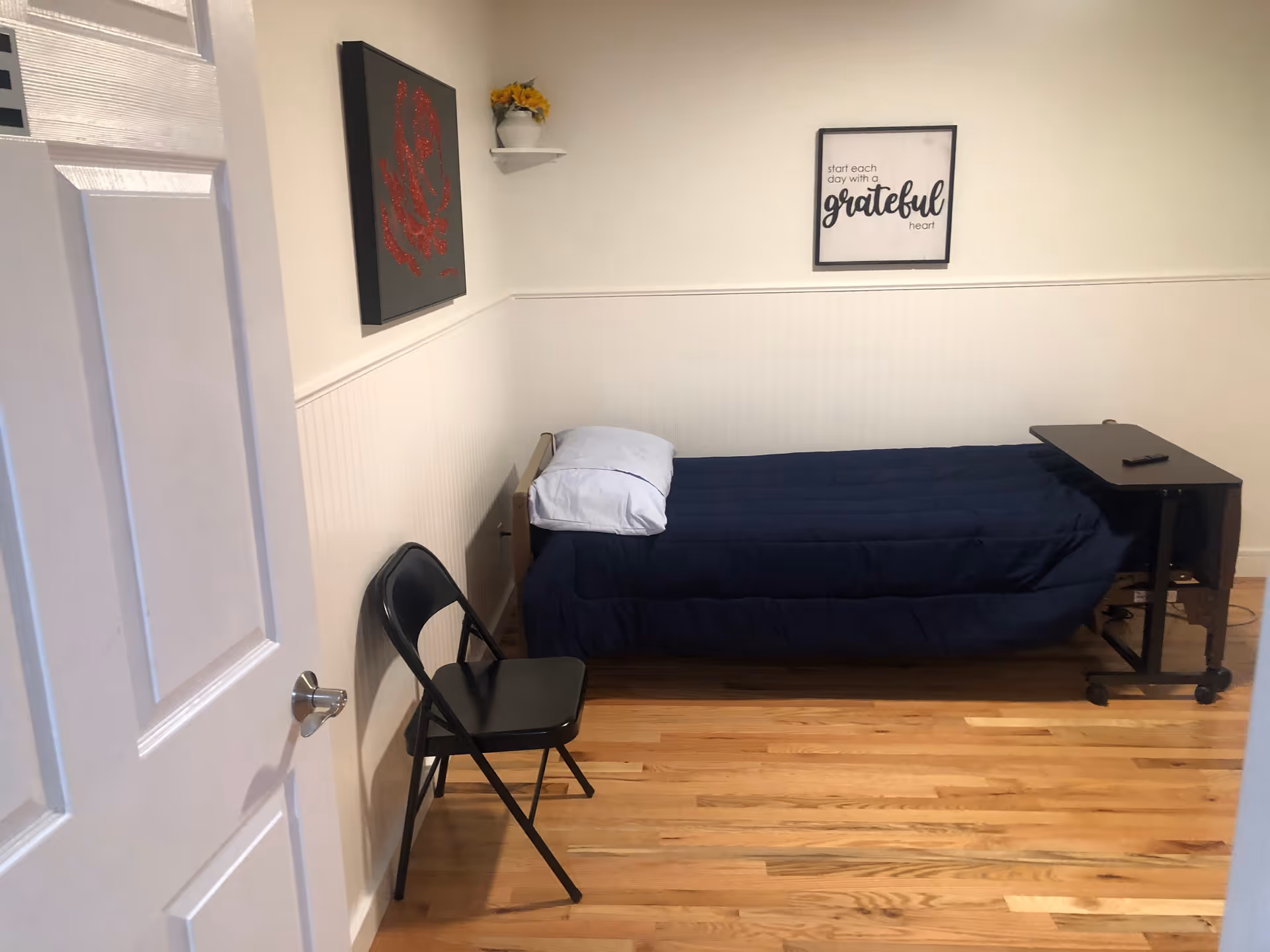 A small bedroom with a single bed covered in a dark blue comforter and a white pillow. Next to the bed is a black folding chair and a small black rolling overbed table with a remote control on it. The walls are white with a white wainscoting halfway up. There is a framed picture on the wall above the bed with the text 'start each day with a grateful heart' and another framed artwork with a red rose on the adjacent wall. The floor is wooden.