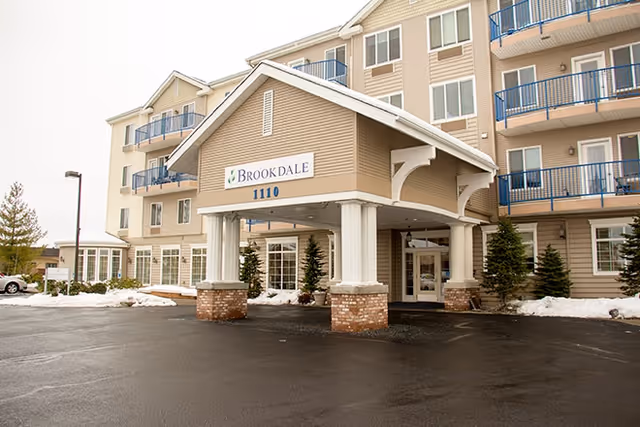 Exterior view of a multi-story senior living facility with beige siding and blue balcony railings. The entrance features a covered porte-cochère supported by white columns with brick bases. Snow is visible on the ground around the building.