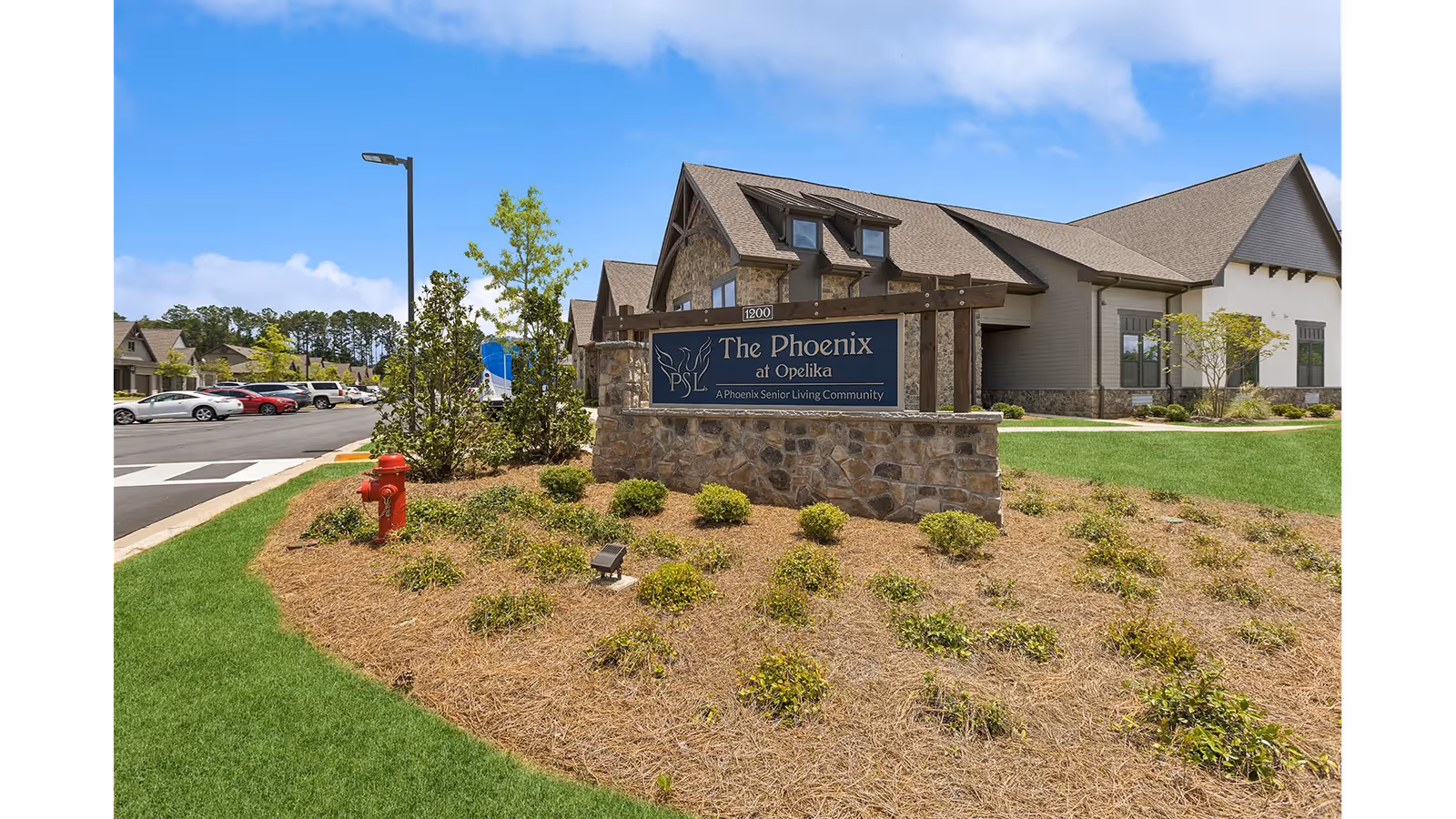 Exterior view of The Phoenix at Opelika senior living community sign surrounded by landscaping with bushes and a fire hydrant, with the building and parking lot visible in the background under a blue sky.