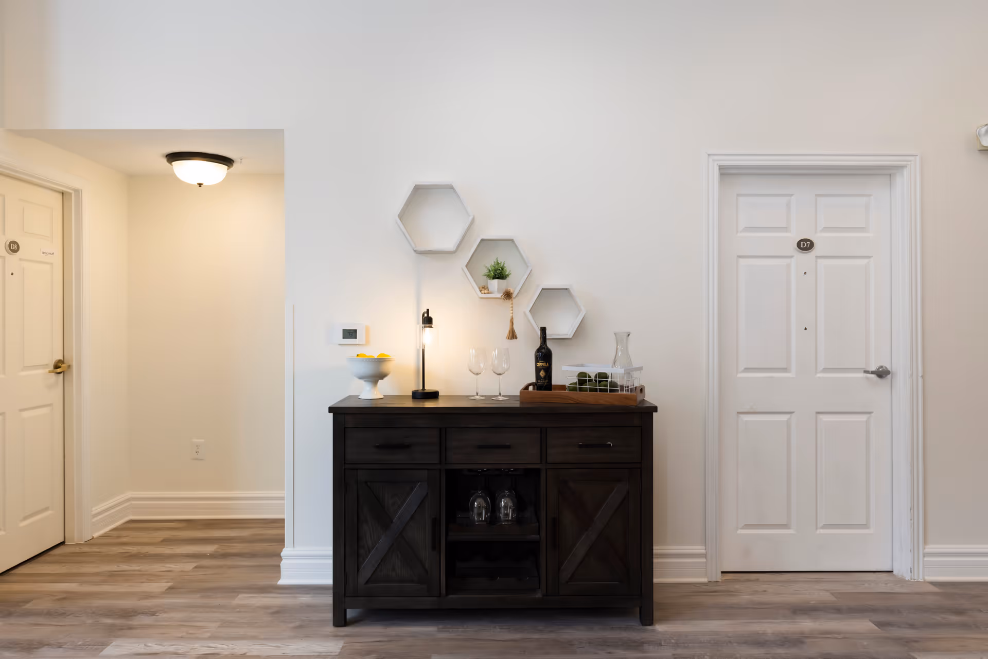 Interior hallway area with two white doors labeled D6 and D7 on either side. Between the doors is a dark wooden sideboard with three drawers and two cabinets. On top of the sideboard are decorative items including a bowl with lemons, a small lamp, two wine glasses, a bottle of wine, a glass pitcher, and a tray with green moss balls. Above the sideboard are three hexagonal wall shelves with small plants and decor. The floor is wood with a light finish and the walls are painted white.