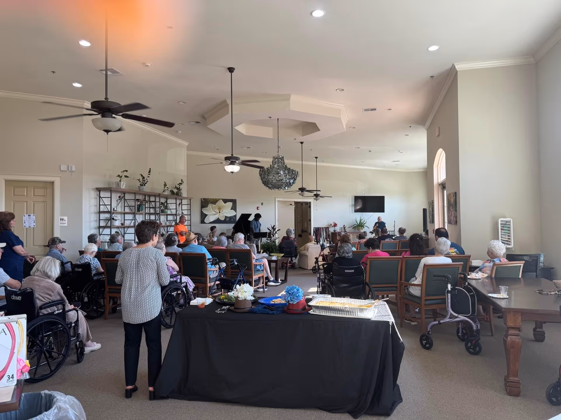 A large communal activity room in a senior living facility with many elderly residents seated and facing a presenter, wheelchairs and walkers, and a buffet table in the foreground.