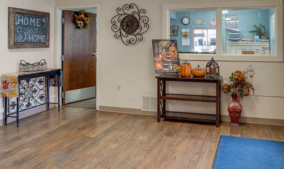 Lobby-style interior with wood flooring, decorative tables, a wall clock and autumn decorations beneath a 'Home Sweet Home' sign and a service window.