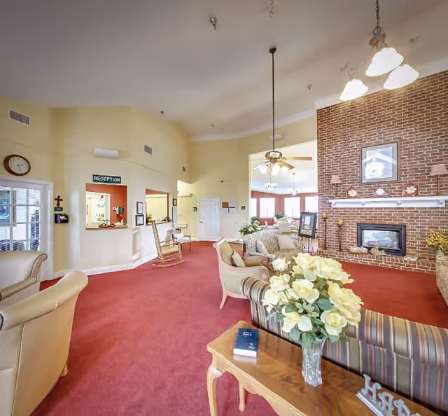 Spacious common lounge with upholstered seating around a brick fireplace, a reception window, and a floral centerpiece on a table.