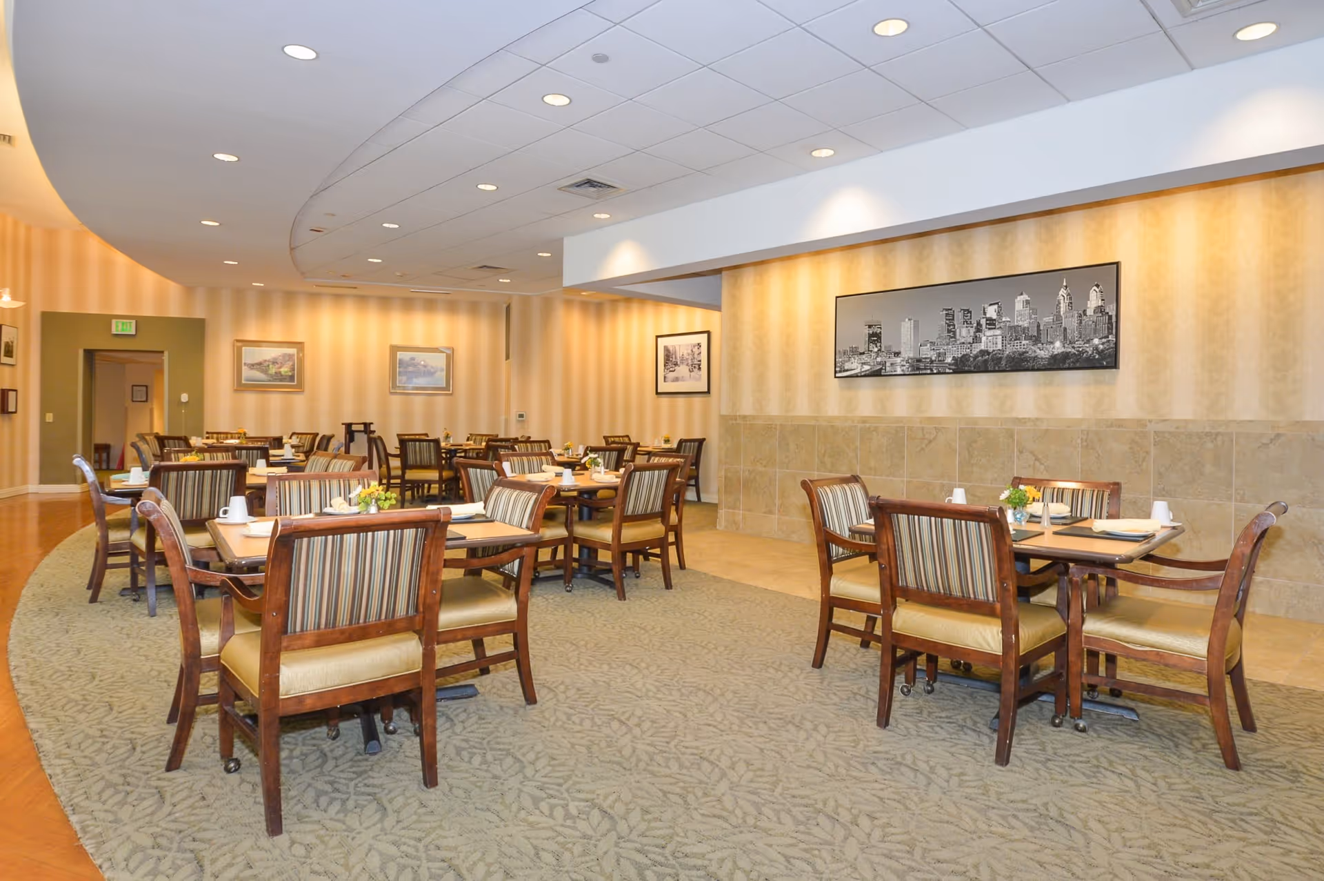 A spacious dining room in a senior living facility with multiple wooden tables and chairs arranged neatly. The chairs have striped upholstery and the tables are set with placemats, napkins, and small flower arrangements. The room features warm beige walls with framed pictures and a large black and white cityscape photograph. The ceiling has recessed lighting and the floor is carpeted with a patterned design.