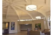 Interior view of a spacious common area in a nursing and rehabilitation center featuring high vaulted ceilings with large circular light fixtures, modern furniture including chairs and sofas, and colorful artwork on the walls.