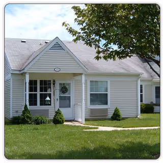 Front exterior of a single-story light-colored townhouse unit with a small covered porch, windows, a tree, and a grassy lawn.