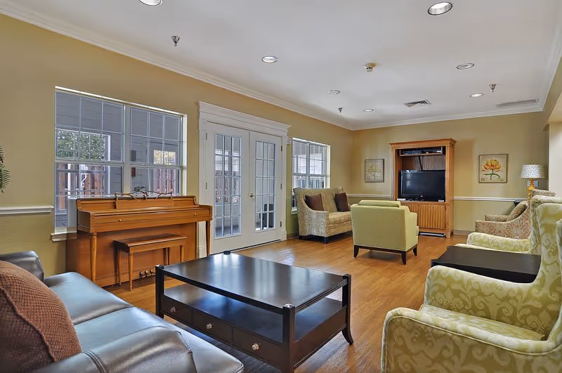 A spacious senior living common area with wooden flooring, beige walls, and recessed ceiling lights. The room features a brown leather sofa with a brown cushion, a black coffee table, two patterned armchairs, a green armchair, a wooden piano with a bench, and a wooden entertainment center with a TV. There are three large windows and a set of French doors letting in natural light.