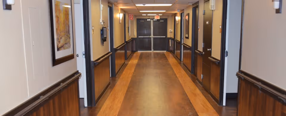 A well-lit hallway in a nursing and rehabilitation facility with wooden paneling on the lower half of the walls, light-colored upper walls, framed artwork, wall-mounted lights, and several closed doors along the corridor leading to double exit doors at the end.