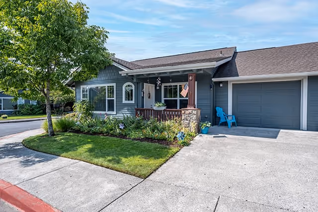 Exterior view of a single-story residential building with a gray facade, a small covered porch with an American flag, a stone pillar, a blue chair near the garage, and a well-maintained lawn with shrubs and a tree.