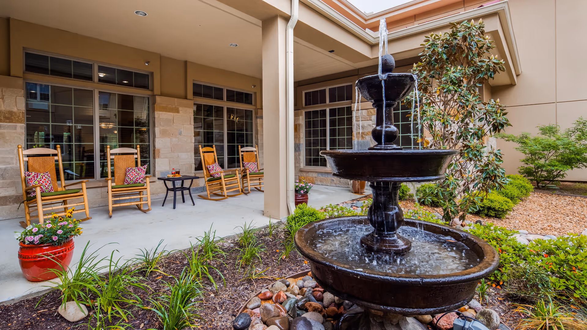 Outdoor patio area at Legend of McKinney featuring a three-tiered water fountain surrounded by landscaped greenery and plants. There are four wooden rocking chairs with colorful cushions arranged along the covered patio next to large windows, with a small table holding drinks between two of the chairs.