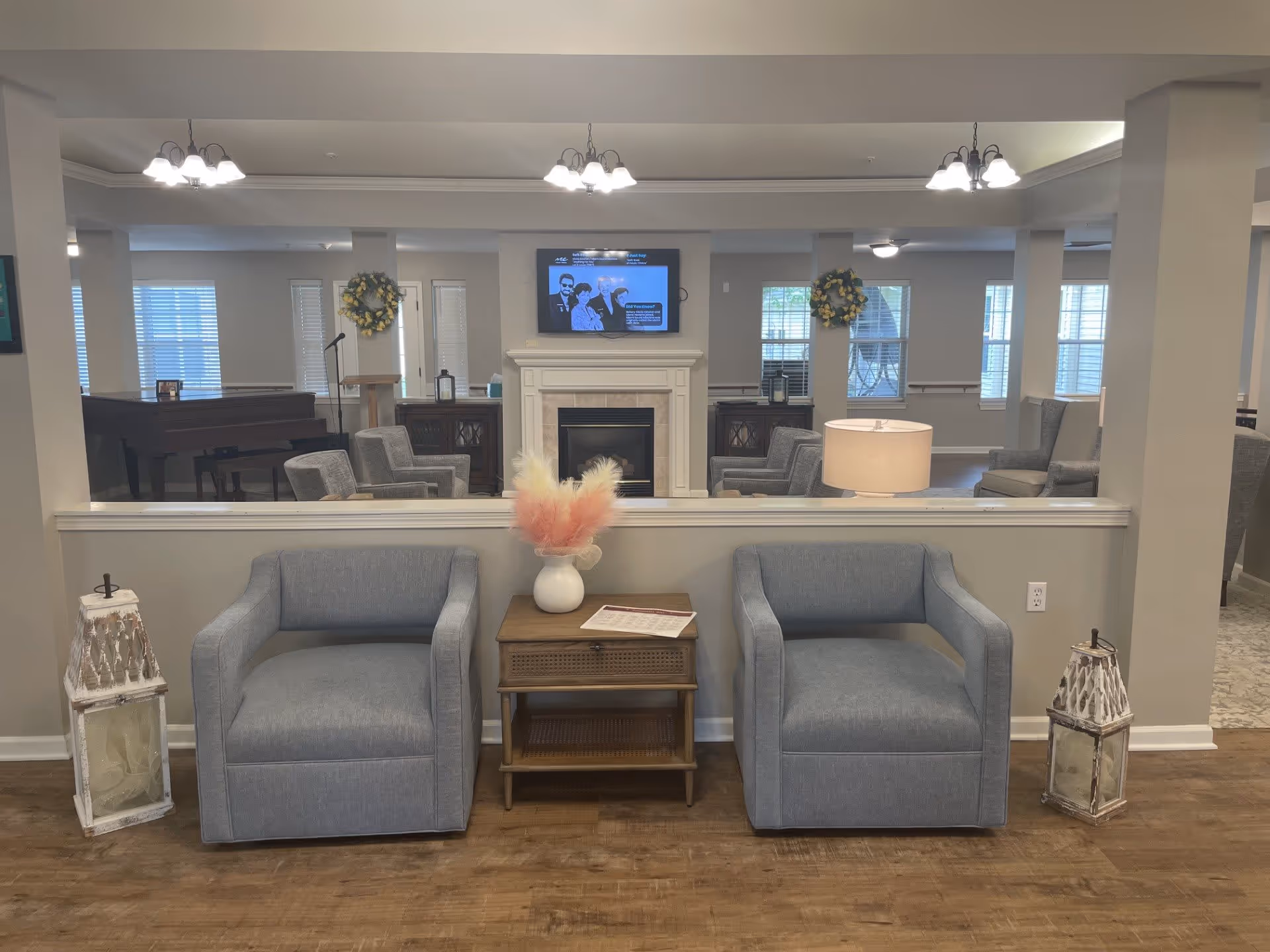 A cozy living room area with two gray armchairs separated by a wooden side table holding a white vase with pink and cream dried flowers. On either side of the chairs are decorative lanterns. In the background, there is a fireplace with a TV mounted above it, flanked by two wreaths and additional seating. The room has soft lighting from ceiling fixtures and a table lamp.