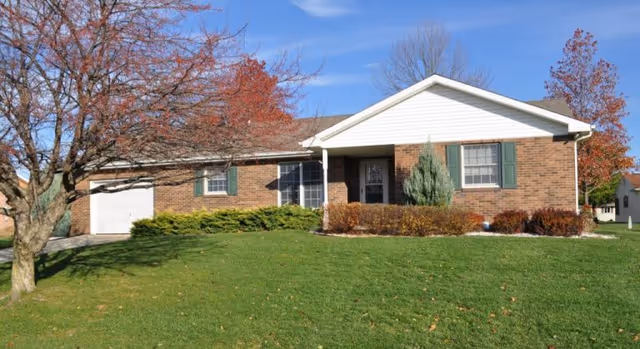 Single-story brick building with a white garage door and green shutters on the windows, surrounded by a well-maintained lawn and trees with autumn foliage under a clear blue sky.