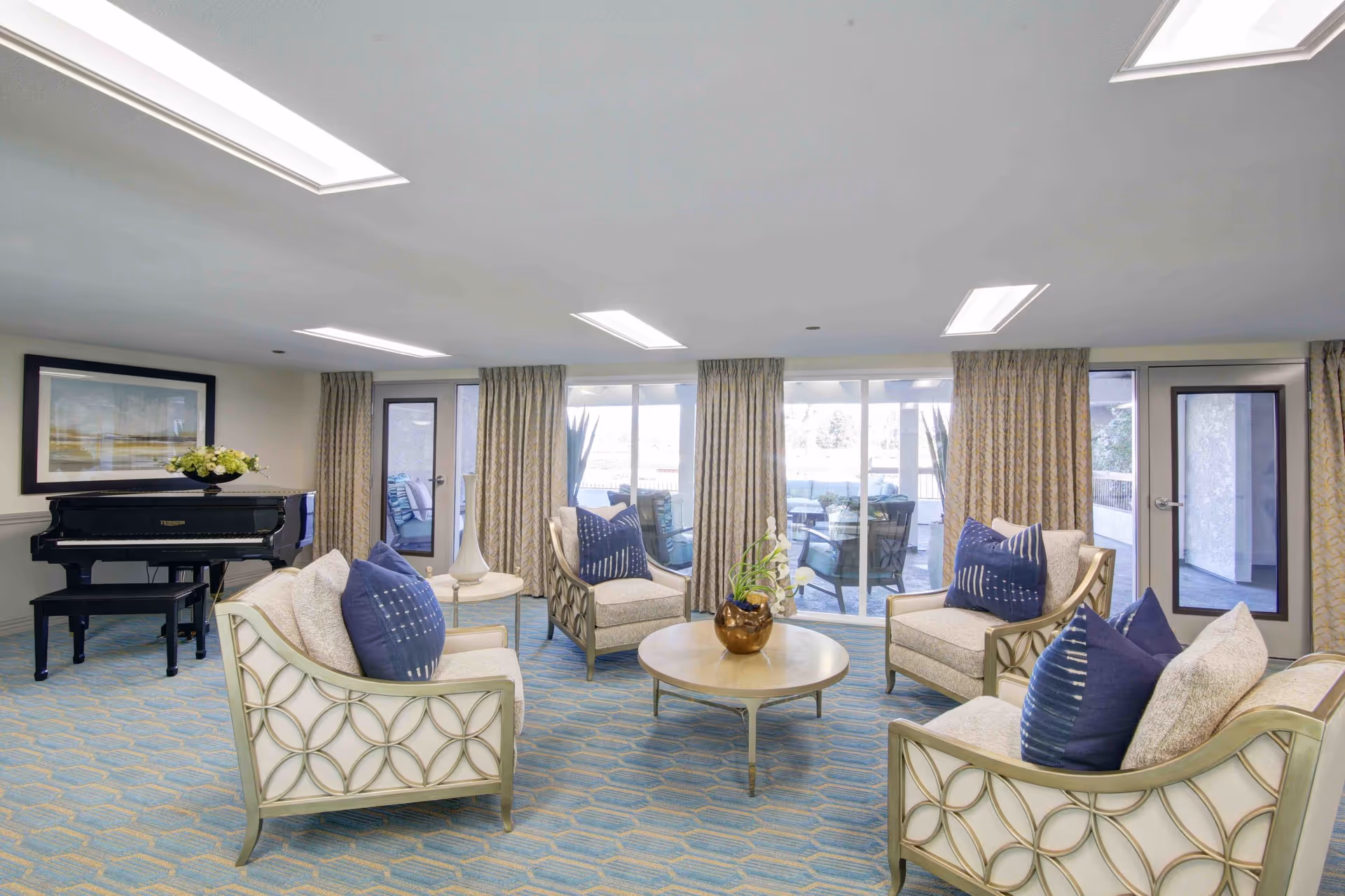 A bright and spacious senior living facility common area with four cushioned armchairs featuring decorative circular patterns on the sides and blue accent pillows. A round wooden coffee table with a bronze vase holding white flowers is centered among the chairs. Large windows and glass doors with beige patterned curtains allow natural light to fill the room. A black grand piano with a floral arrangement on top is positioned against the wall, beneath a framed landscape painting. The room has a light blue patterned carpet and recessed ceiling lights.