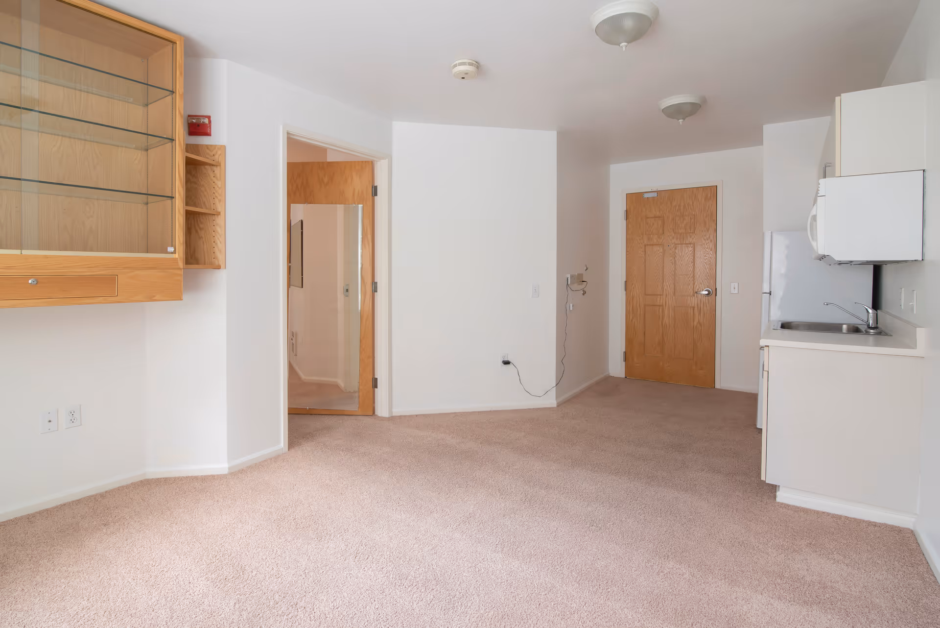 Empty room with beige carpet, white walls, and wooden doors. There is a small kitchenette on the right side with a sink, microwave, and refrigerator. On the left side, there is a wooden cabinet with glass shelves mounted on the wall. The room is well-lit with ceiling lights.