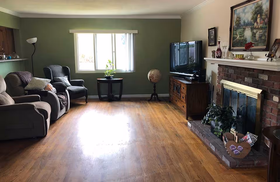 A cozy living room with wooden flooring, a green accent wall with a window, two armchairs and a recliner on the left side, a wooden TV stand with a flat-screen TV on the right, and a brick fireplace with a mantel decorated with various items including a painting, vases, and candles. There is also a globe on a stand and a small table with a plant near the window.