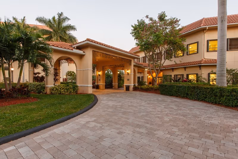 Exterior view of Oakbridge Terrace Assisted Living Residence at Edgewater at Boca Pointe showing a paved driveway leading to a covered entrance with beige walls and terracotta roof tiles, surrounded by palm trees and landscaped greenery.