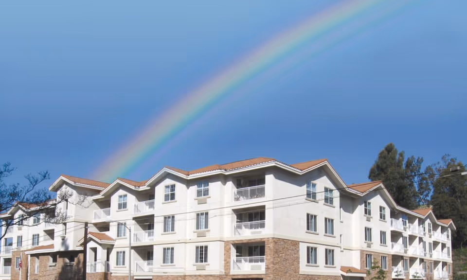 A multi-story residential building with white walls and brown tiled roofs under a clear blue sky with a vibrant rainbow arching above it. Trees and power lines are visible around the building.