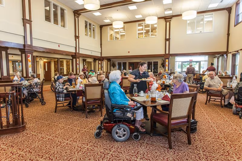 A spacious dining room in an assisted living facility with elderly residents seated at tables, some in wheelchairs, enjoying a meal. A staff member is serving food to one of the tables. The room has high ceilings, large windows, and warm lighting.