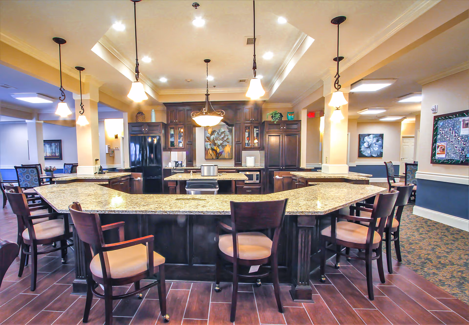 A spacious kitchen area with a large granite countertop island surrounded by wooden chairs with cushioned seats. The kitchen features dark wood cabinetry, a black refrigerator, a microwave, and pendant lighting hanging from the ceiling. In the background, there are dining tables and chairs, artwork on the walls, and a bulletin board.